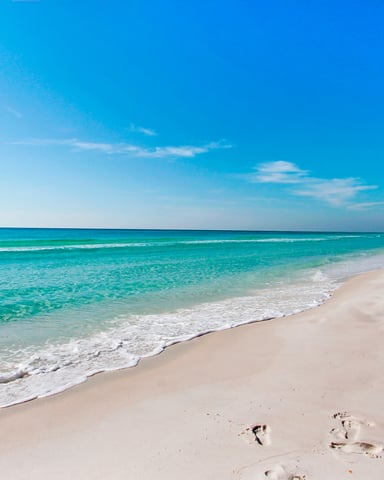 Sandy beach with turquoise water, white waves, and high-rise buildings along the shoreline under clear blue sky