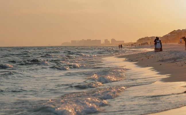 Sandy beach at sunset with rolling waves, beachgoers, and high-rise buildings along the shoreline in the distance.